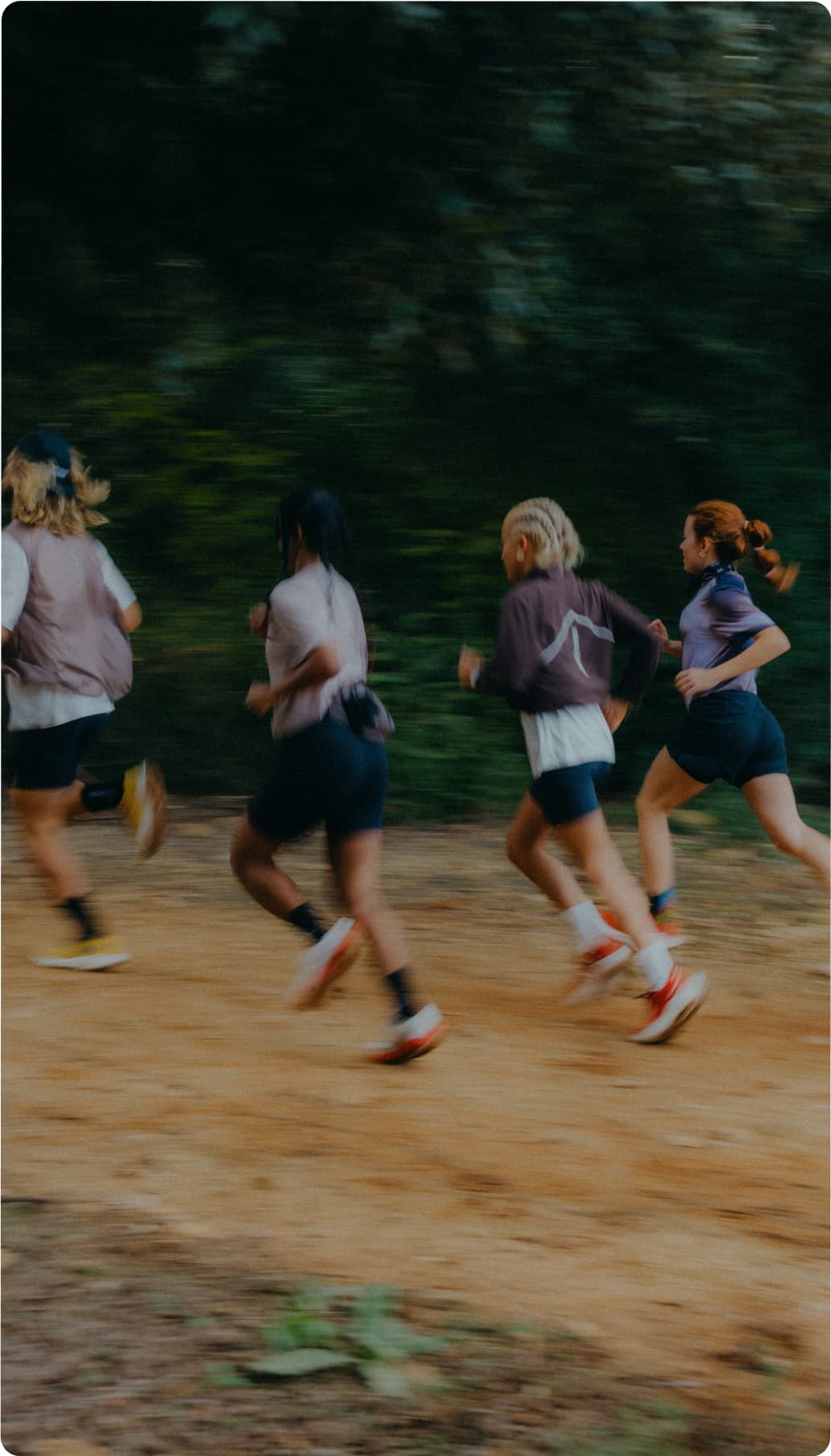 Four people running on a dirt path in a forested area, wearing athletic gear, with blurred motion indicating speed.