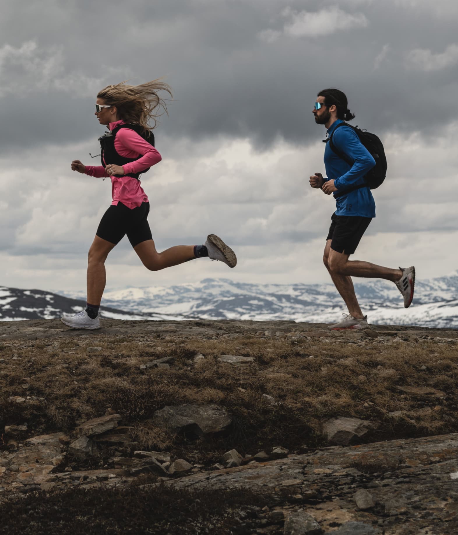 Two people running on a mountain trail, wearing athletic gear, under a cloudy sky with snow-dusted peaks in the background.