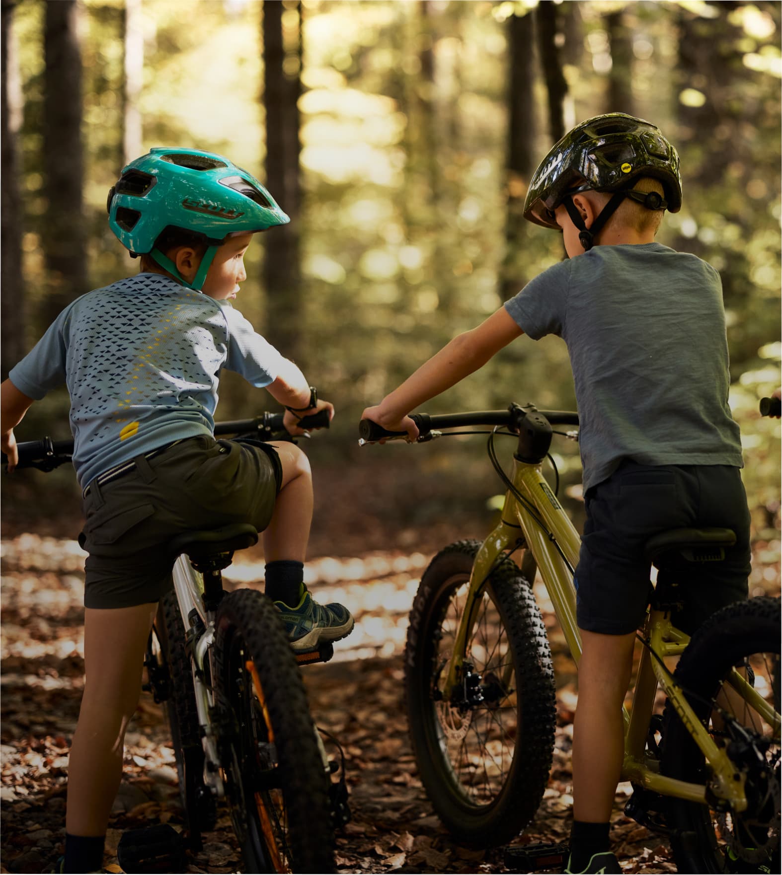 Two children riding bicycles on a forest trail, surrounded by trees and dappled sunlight.