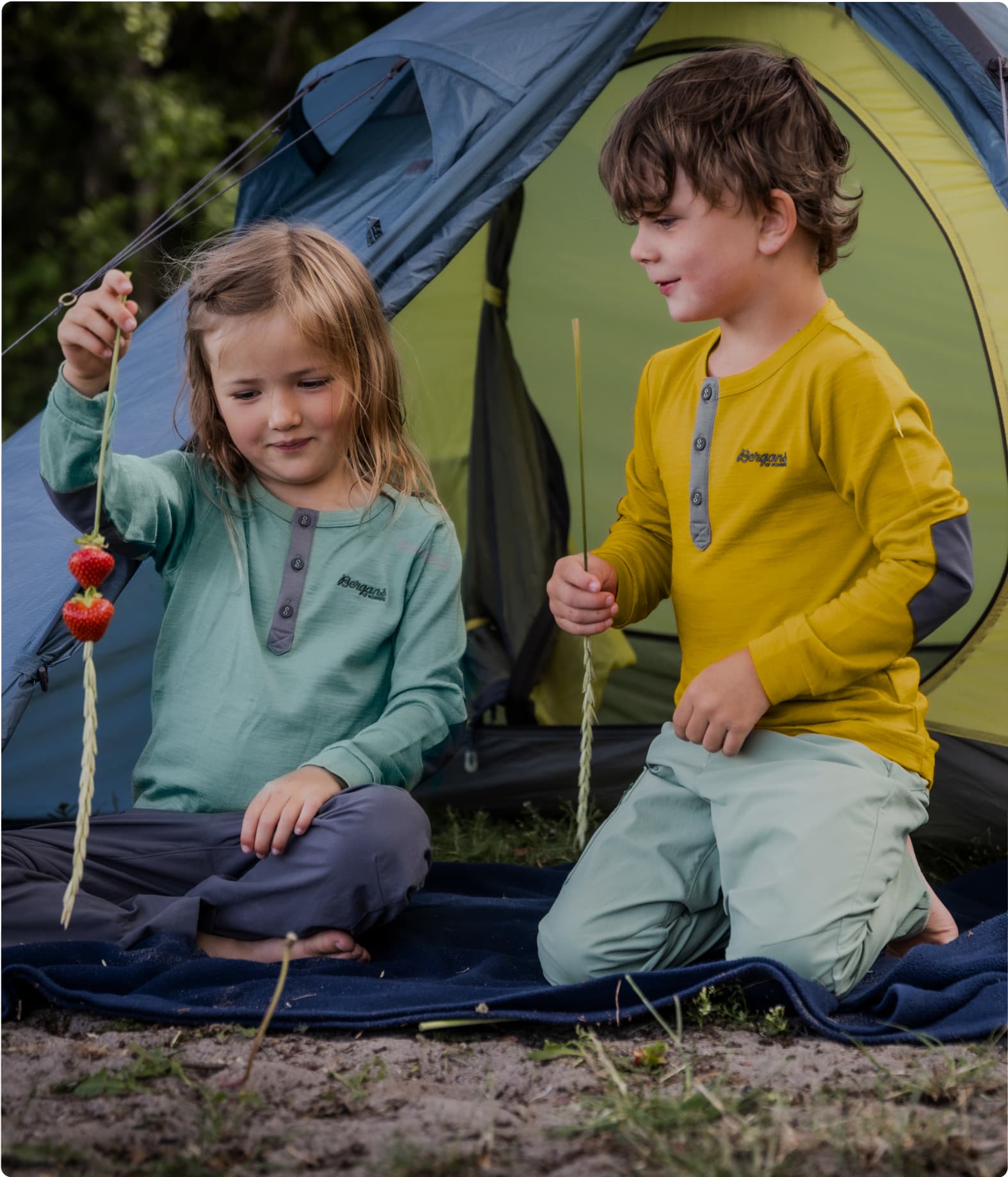 Two children in colorful shirts sit at a tent entrance, one is holding a straw with strawberries.