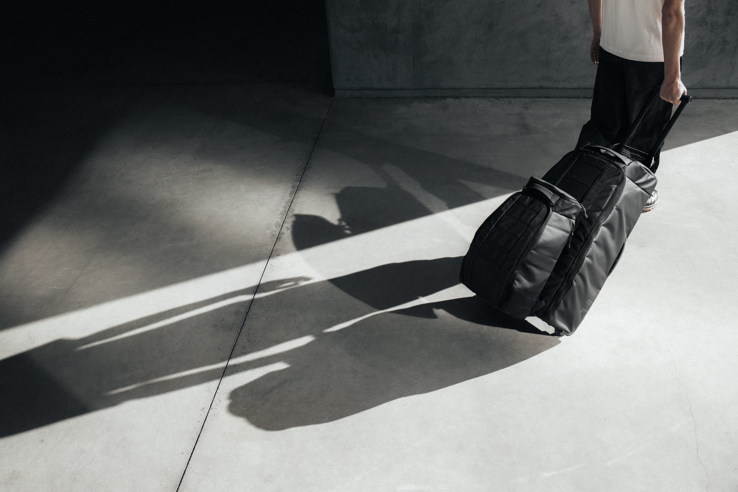 Person pulling a large black suitcase on wheels across a sunlit concrete floor, casting a shadow.
